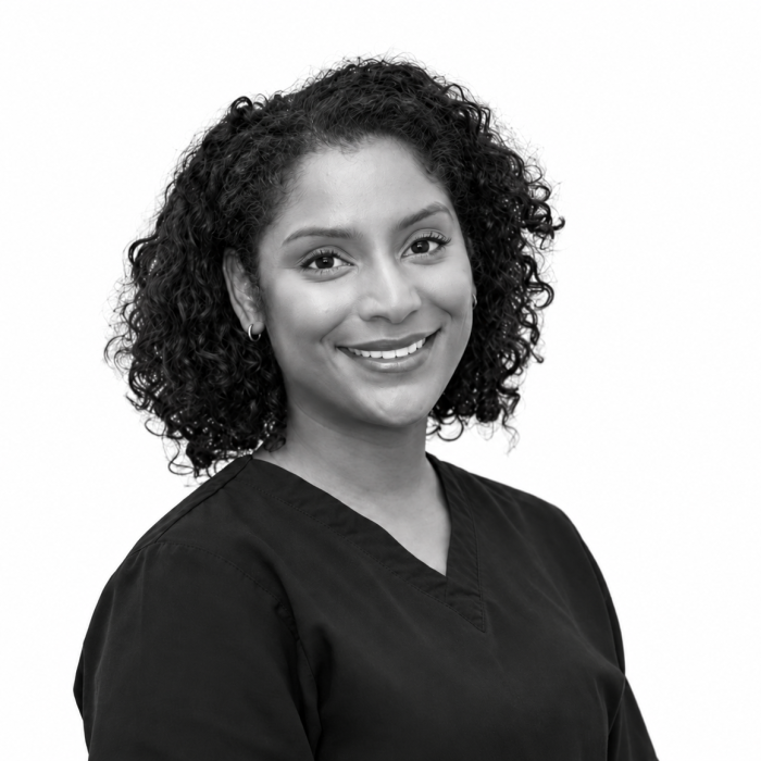 Black and white portrait of Dr Amelia Simone Bhoorasingh, general and restorative dentist at 72 Dental, smiling in black scrubs against a white background